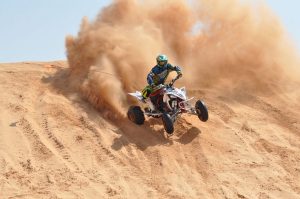 An ATV racer jumps off a sandy hill kicking up dust behind him