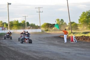 Three ATVs race on a dirt track while the official waves a green flag