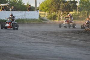 Three ATVs race on a dirt track with trees and spectators in the distance