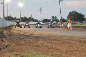 Four ATVs race on a dirt track with flood lights in the distance