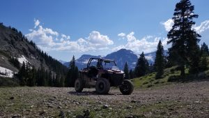 A UTV sits on a dirt road with mountains and evergreen trees behind it