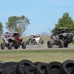 Three UTVs including the JSR Performance UTV number 911 race around the curve of the asphalt track with a wall of protective tires on the side of the track