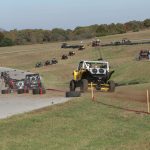A yellow UTV is following a line of other UTVs on a dirt track during a competition